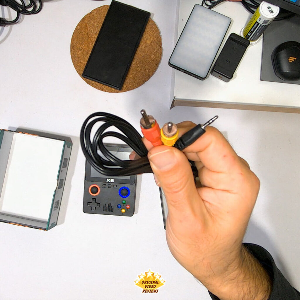 Hand holding an AV cable with yellow video, red audio, and black 3.5mm connectors, with the X6 handheld console and packaging visible in the background.