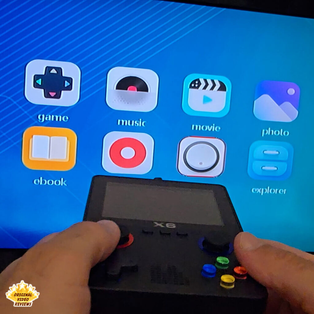 Hands holding the X6 handheld console connected to a TV, showing the main menu with game, music, movie, photo, ebook, and explorer icons.