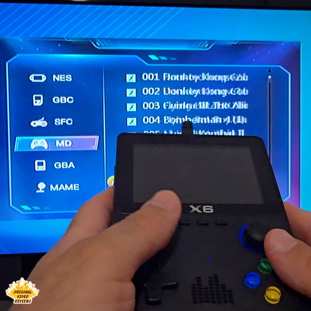 Hands holding the X6 handheld console while navigating its menu on a TV screen, showing NES, GBC, SFC, MD, GBA, and MAME emulator options.