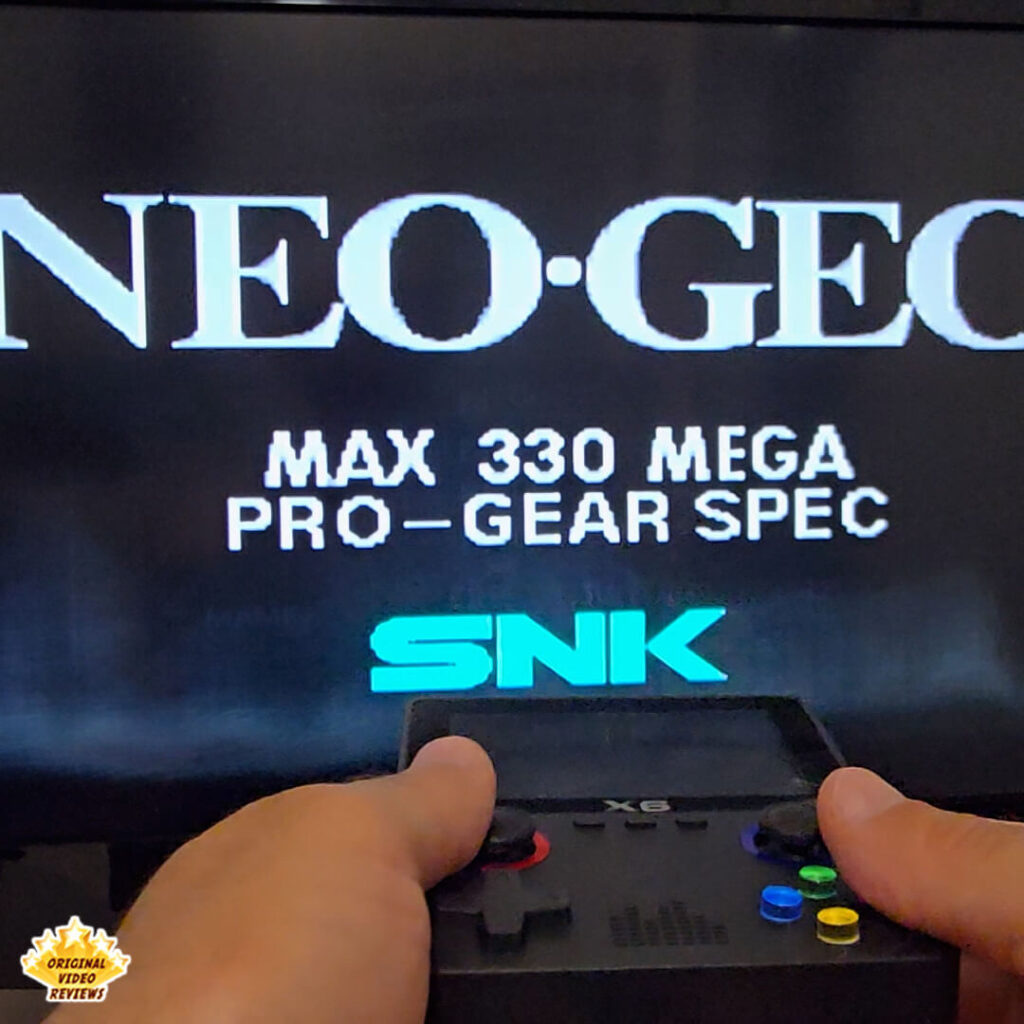 Hands holding the X6 handheld console connected to a TV displaying the Neo Geo Max 330 Mega Pro-Gear Spec startup screen, with the OVR Original Video Reviews logo.