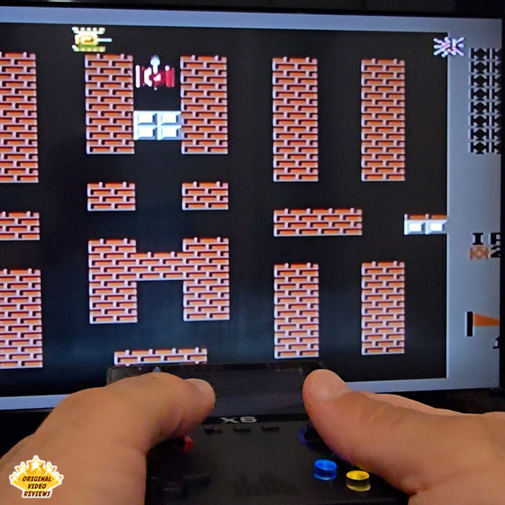 Close-up of hands holding the X6 console while playing the classic NES game "Battle City" on a TV screen.