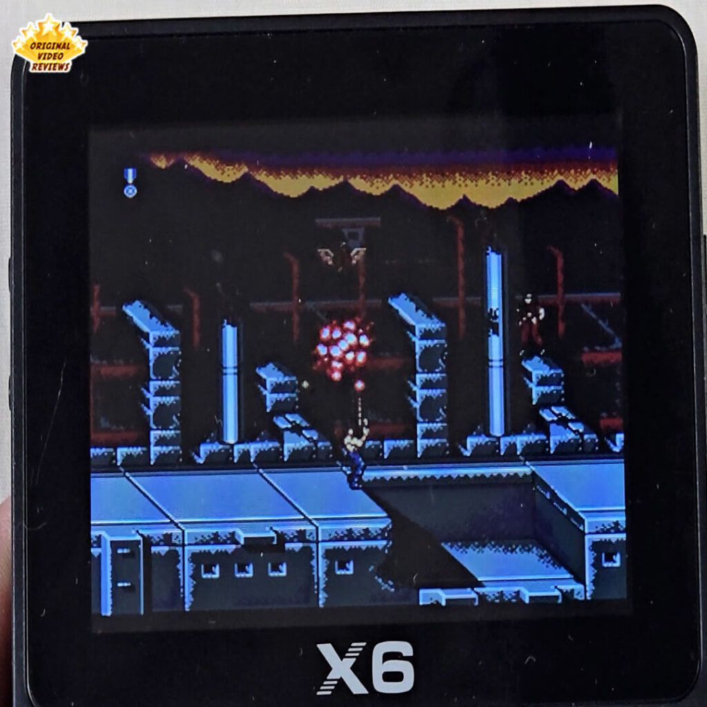 Gameplay screenshot from Contra displayed on the X6 handheld console, showing the player shooting enemies in a destroyed futuristic environment.