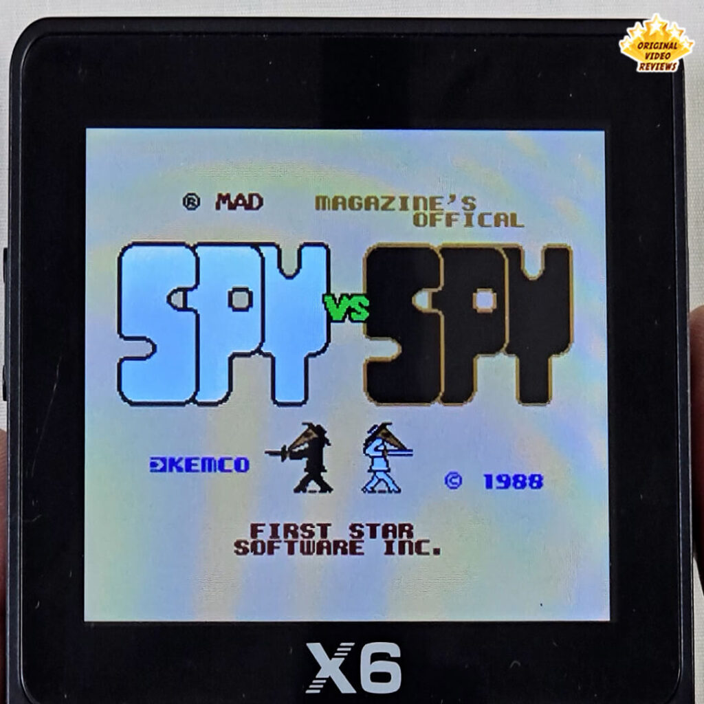 Title screen of Spy vs Spy displayed on the X6 handheld console, showing the black and white spies from MAD Magazine.