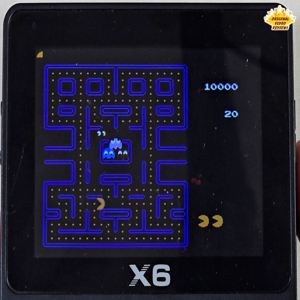 X6 handheld console showing Pac-Man gameplay with the character navigating a maze while being chased by blue ghosts.