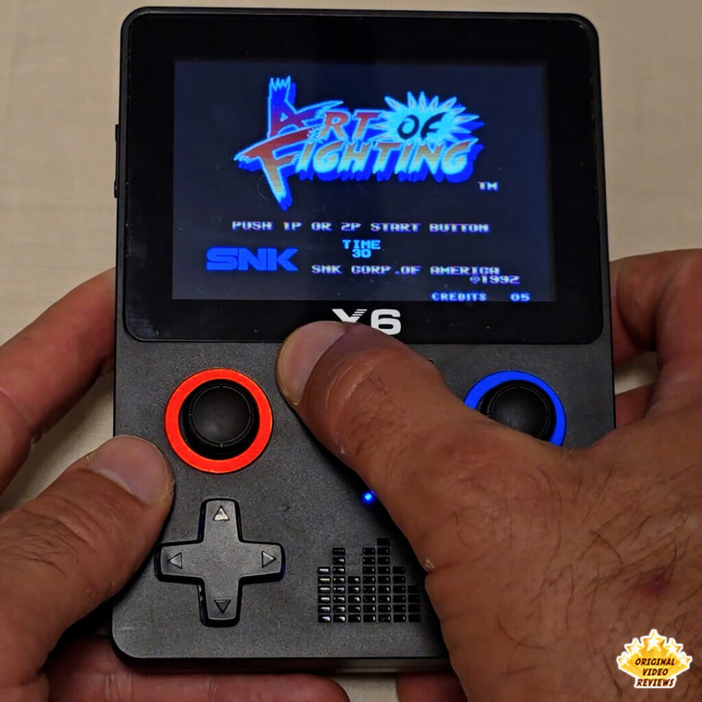 Close-up of a person holding an X6 handheld gaming console playing the game "Art of Fighting" by SNK. The title screen of the game is visible on the console's screen.