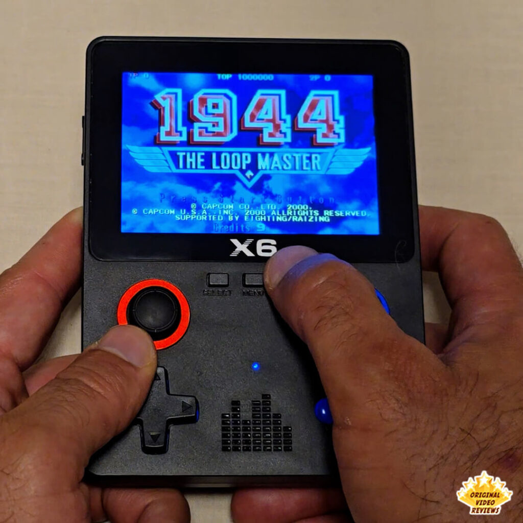 Close-up of the X6 handheld retro gaming console displaying the title screen of "1944: The Loop Master". The console's buttons and joysticks are clearly visible.