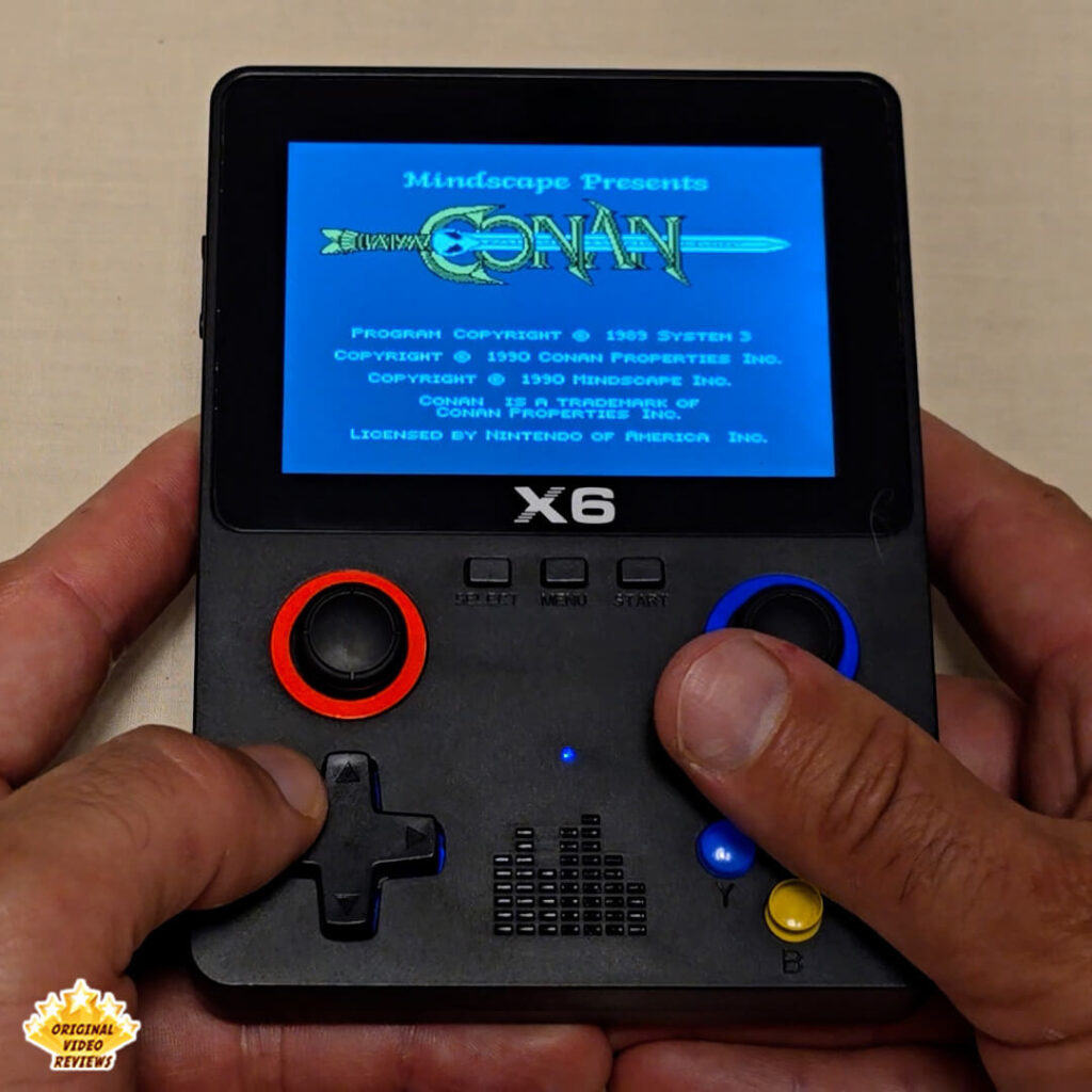 Close-up of the X6 handheld retro gaming console displaying the title screen of "Conan". The console's controls and logo are visible while the game screen shows blue text and graphics.