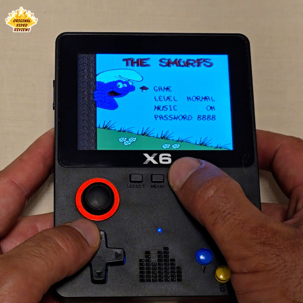 Close-up of the X6 handheld retro gaming console displaying the game menu screen of "The Smurfs". The console's buttons and joysticks are clearly visible while a player navigates the menu options.