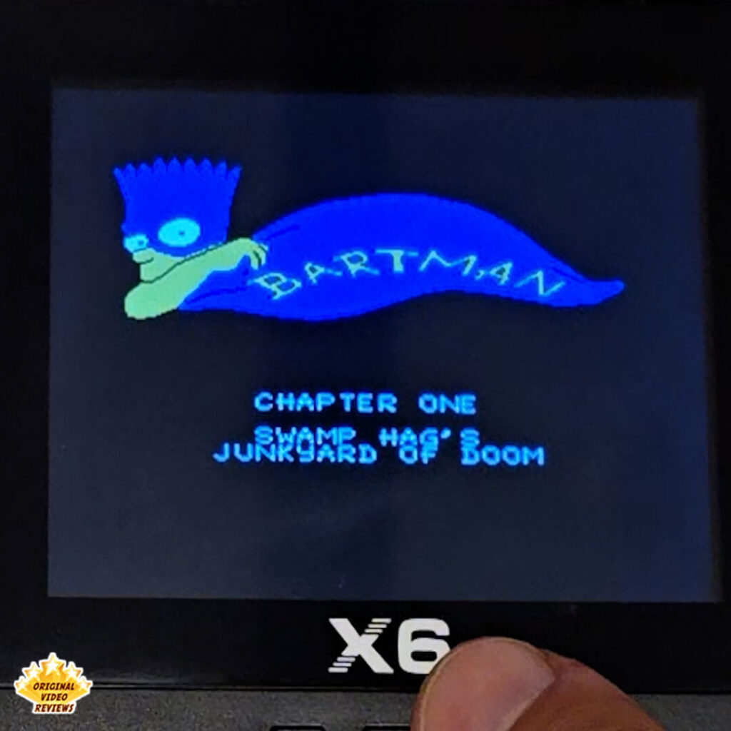 Close-up of the X6 handheld retro gaming console showing Bartman from The Simpsons game on the title screen with "Chapter One - Swamp Hag's Junkyard of Doom". The console's buttons and logo are visible.