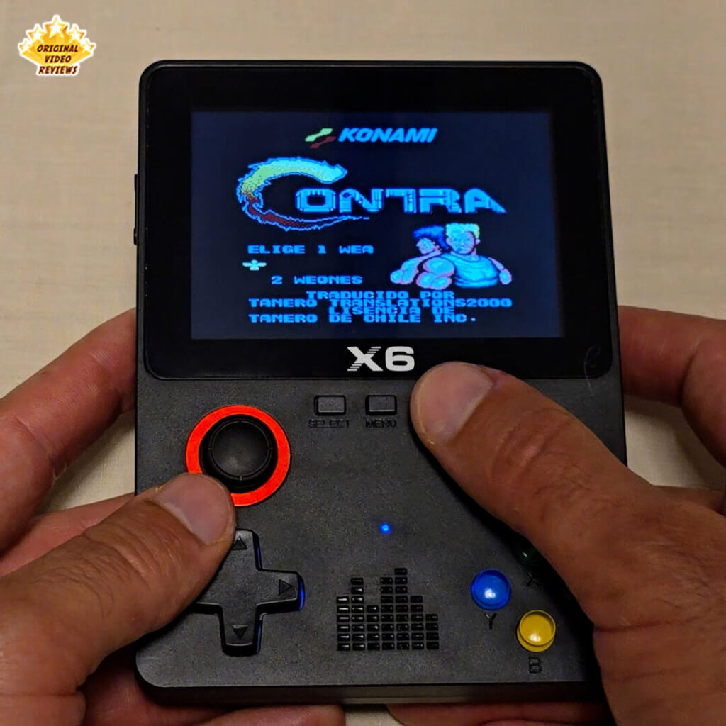 Close-up of the X6 handheld retro gaming console displaying the title screen of Contra by Konami. The screen shows the game's logo and character selection menu in Spanish.