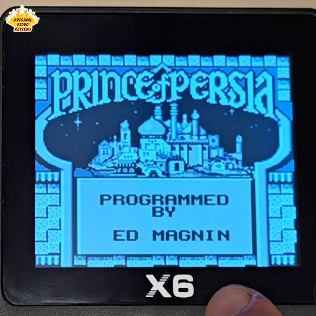 Close-up of the X6 handheld retro gaming console displaying the title screen of Prince of Persia. The console's screen shows the iconic cityscape and credits.