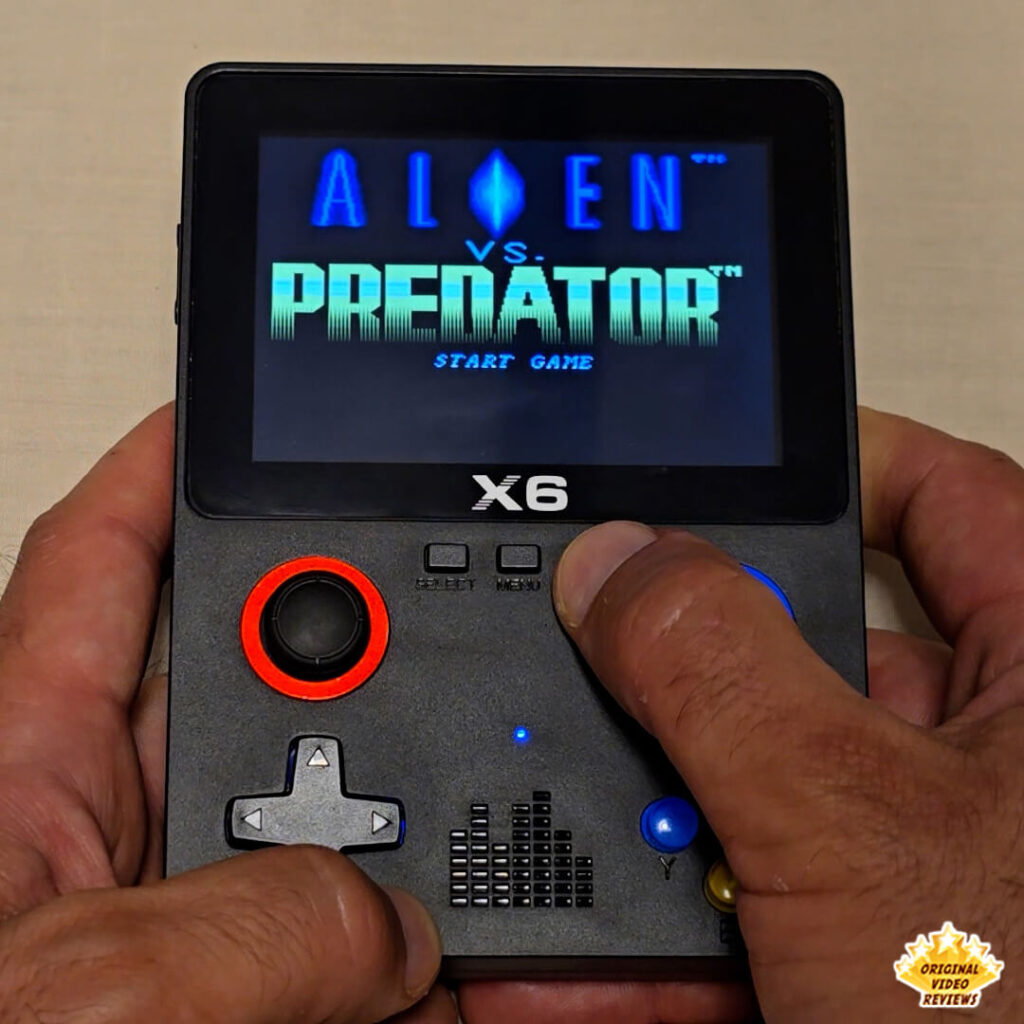 Close-up of the X6 handheld retro gaming console displaying the start screen of Alien vs. Predator. The console's buttons and joysticks are clearly visible.