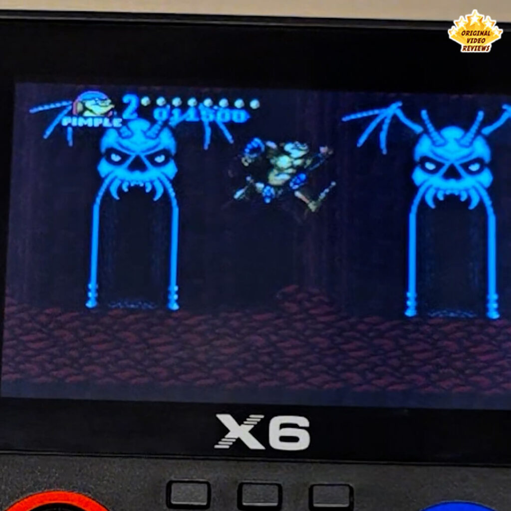 Close-up of the X6 handheld retro gaming console displaying gameplay from "Battletoads," showing the character Pimple mid-jump in a dark cave environment.