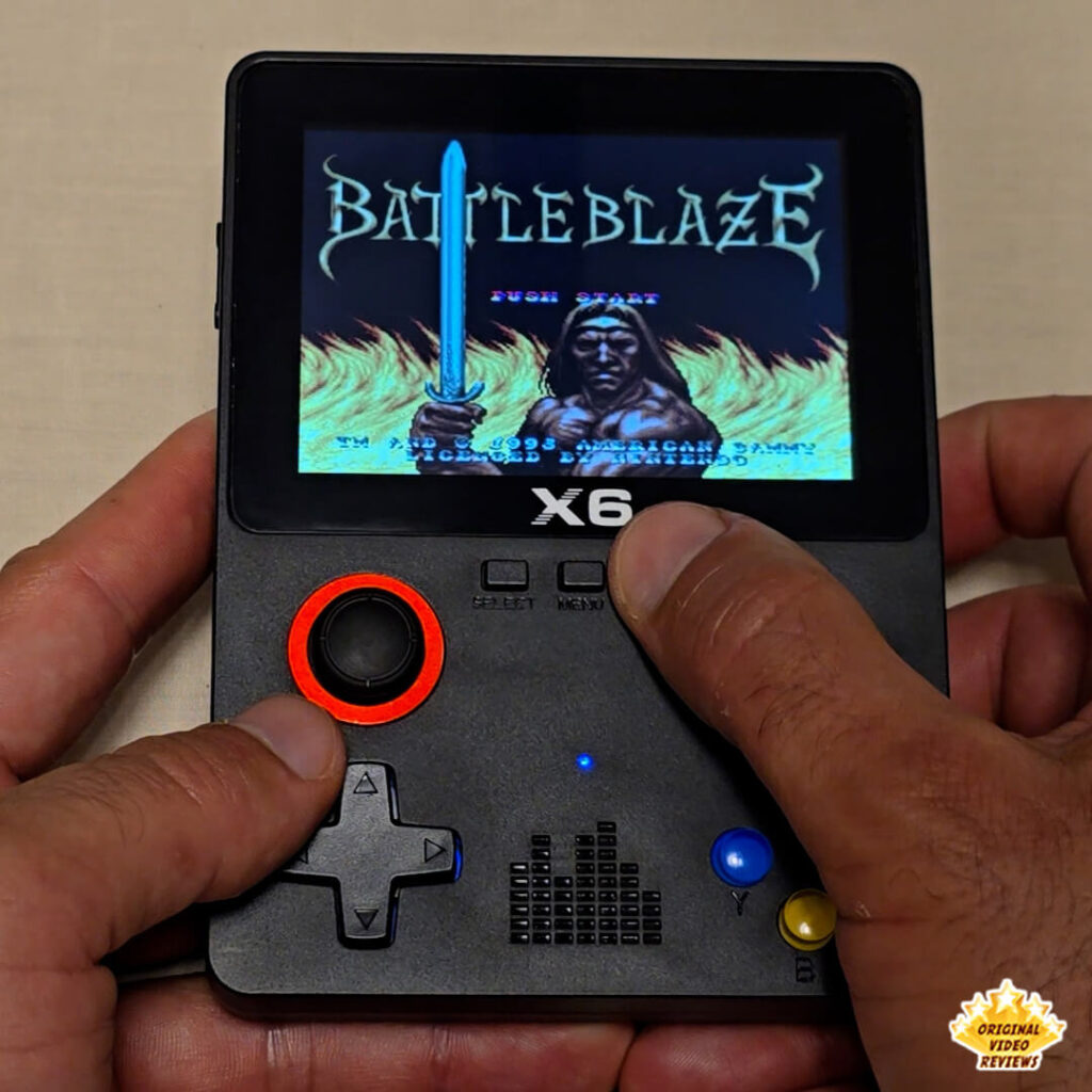Close-up of the X6 handheld retro gaming console displaying the title screen of "Battle Blaze," showing a warrior holding a sword with flames in the background.