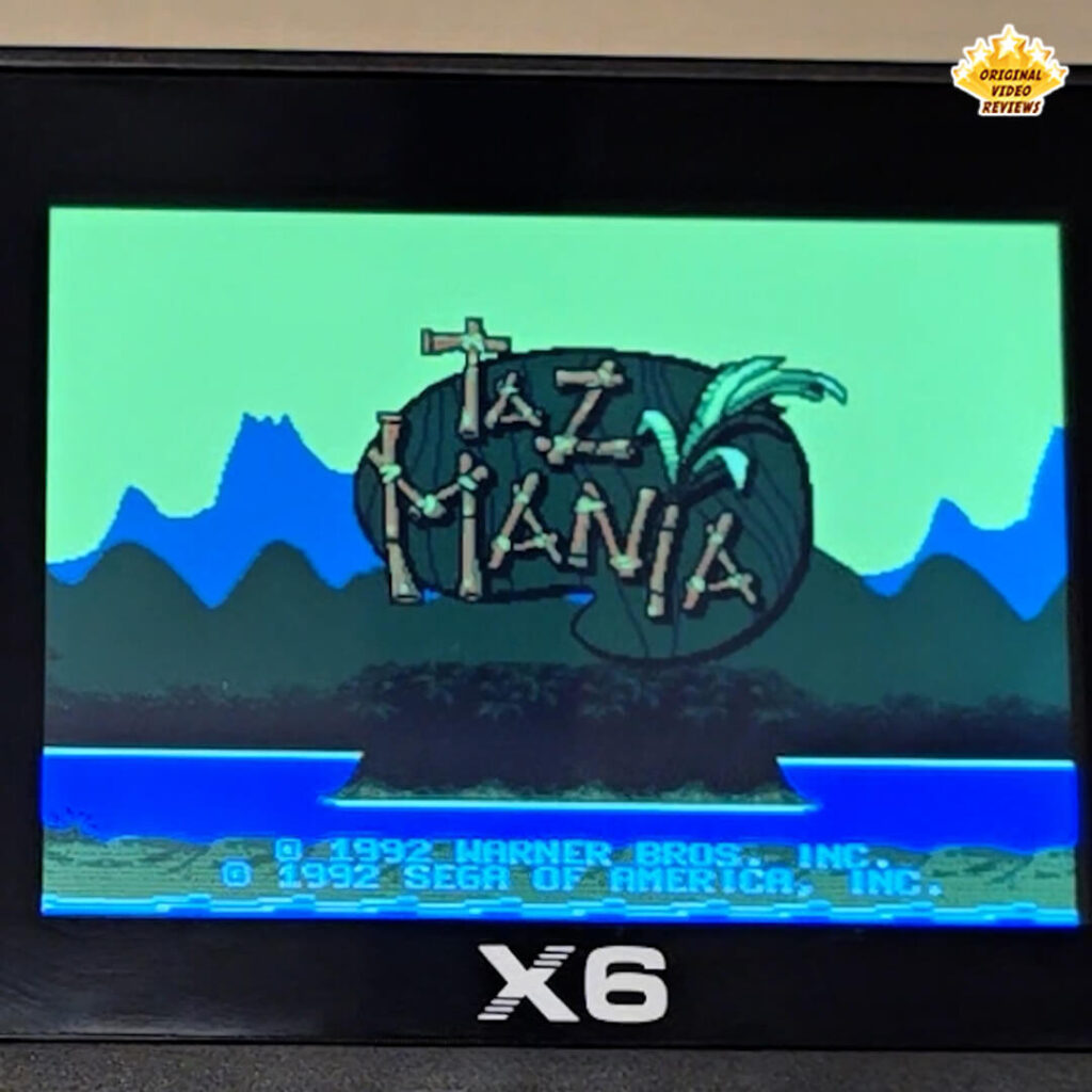 Close-up of the X6 handheld retro gaming console displaying the title screen of "Taz-Mania," showing the game's logo with a tropical background.