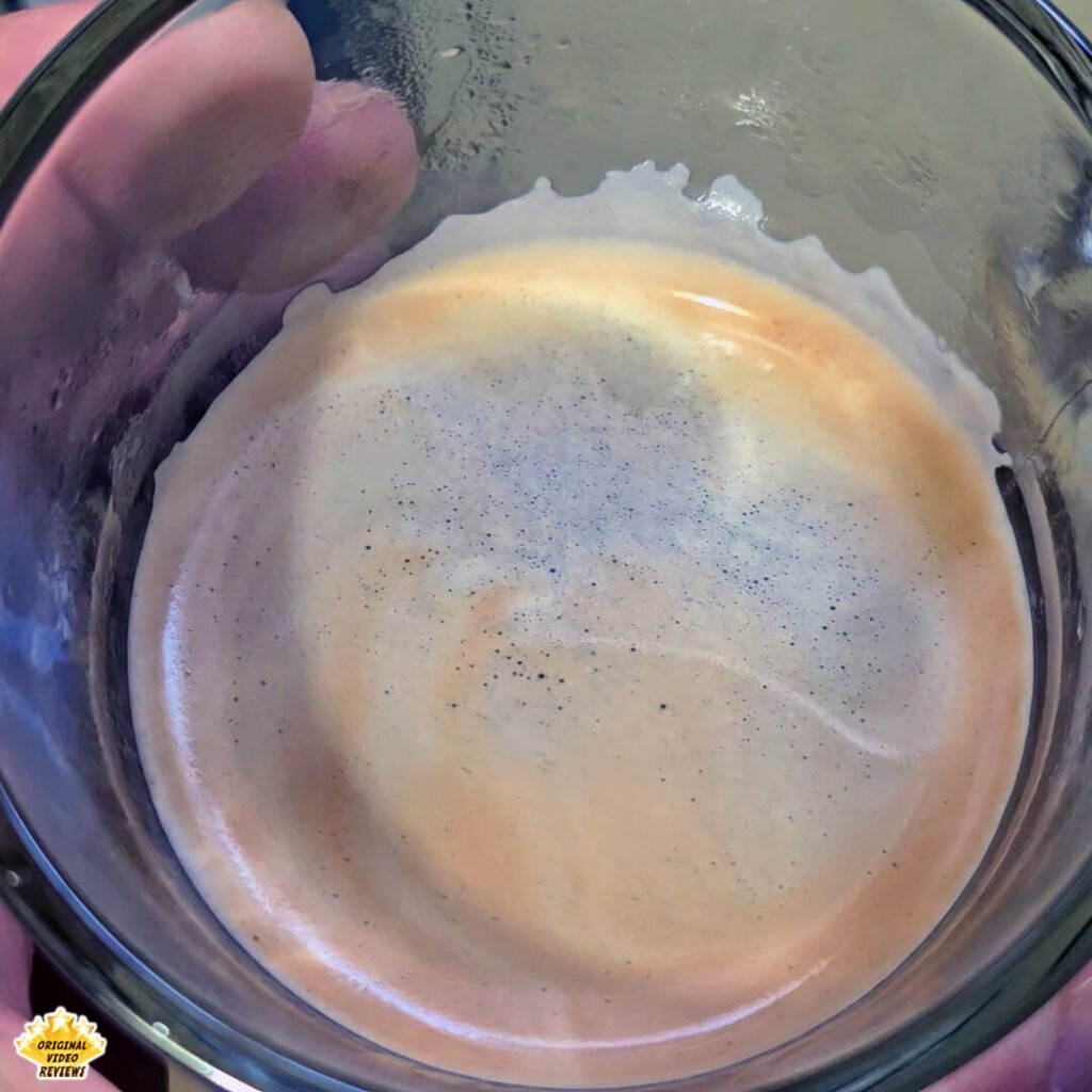 Top-down view of a glass mug showing the crema layer of a freshly brewed Americano made with Bristot Classico Oro coffee beans. The OVR Guy highlights the color, texture, and light bubbles formed on the crema during the first brewing test.
