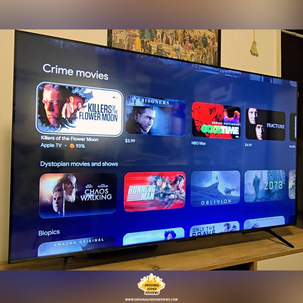 Angled view of the TCL P89K 65-inch 4K QLED TV screen turned on, showcasing the Google TV interface with movie recommendations like "Killers of the Flower Moon," reviewed by Original Video Reviews.