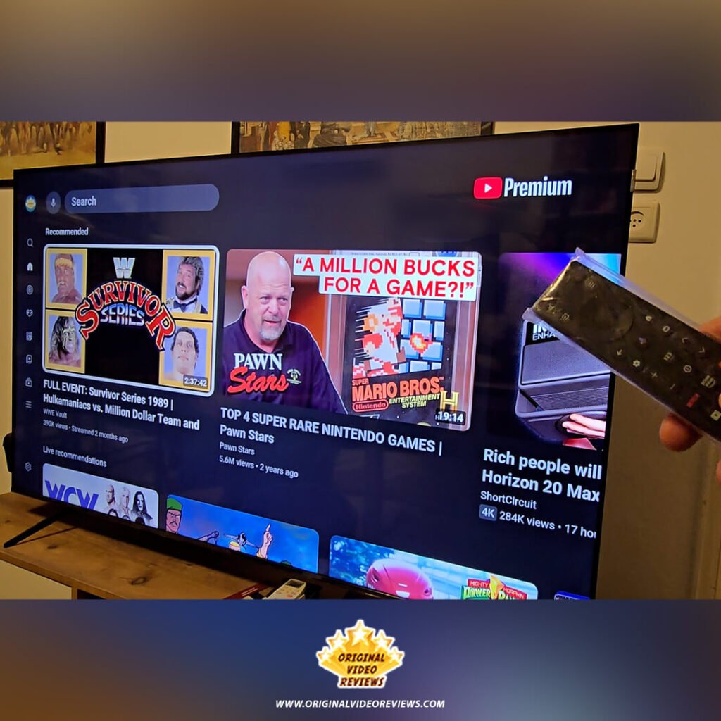 The YouTube Premium interface on the TCL P89K 65-inch 4K QLED TV, showing recommendations for Pawn Stars and ShortCircuit, with the remote visible in hand, from a review by Original Video Reviews.