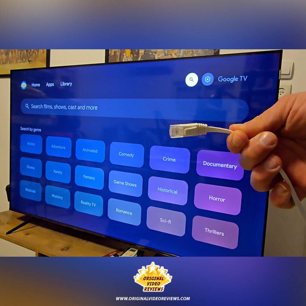 The OVR Guy holding a white Ethernet (LAN) cable in front of the TCL P89K 65-inch 4K QLED TV, comparing wired vs. wireless connectivity for stable 4K streaming during a review by Original Video Reviews.