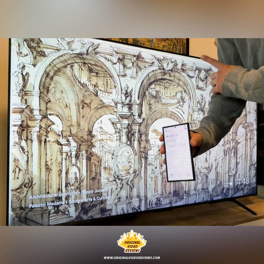 The OVR Guy holding a smartphone with the TCL Home app open, demonstrating the TCL P89K 65-inch QLED TV's role as a smart home hub while the screen is in screensaver mode.