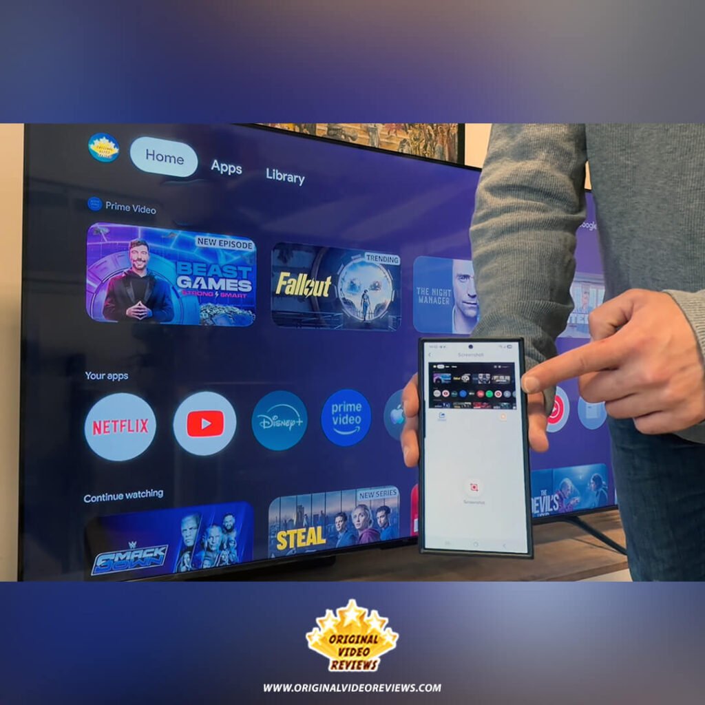 The OVR Guy holding a smartphone and pointing at the screenshot feature in the app, with the TCL P89K 65-inch QLED TV home screen in the background, from a review by Original Video Reviews.