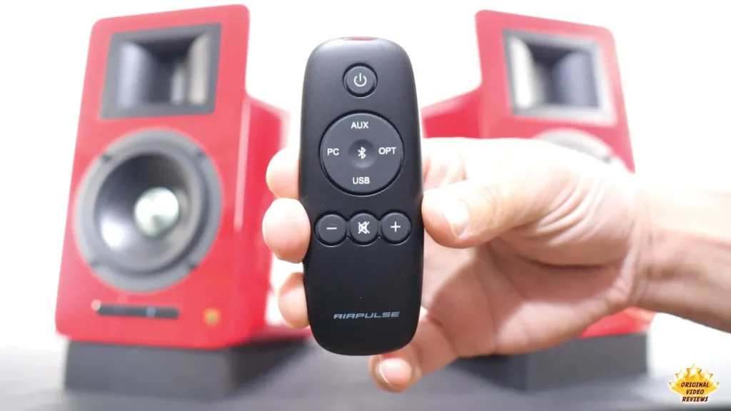 Hand holding the AIRPULSE A100 remote control in front of the speakers