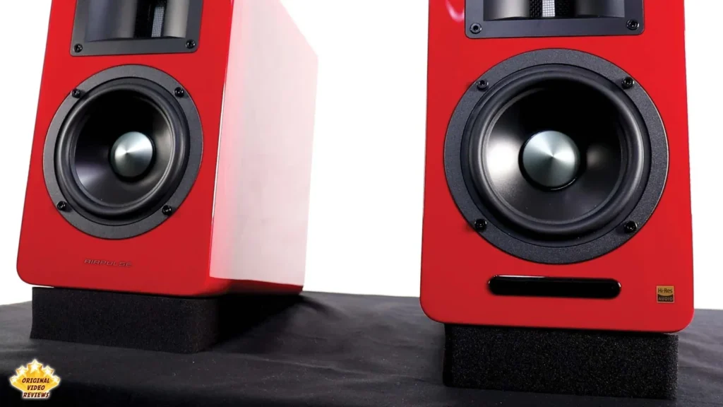 Two red AIRPULSE A100 speakers on a table, one showing its woofer and the other its front port