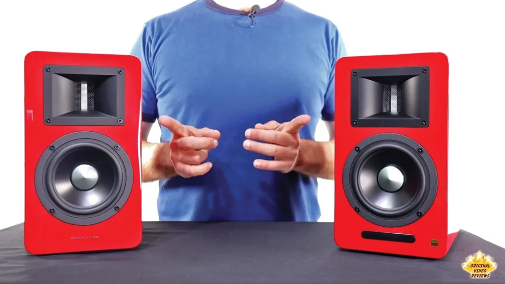 Reviewer gesturing between two red AIRPULSE A100 speakers on a table