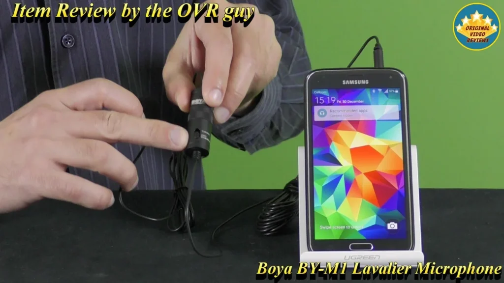 Holding the BOYA BY-M1 Lavalier Microphone control capsule next to a Samsung smartphone on a stand