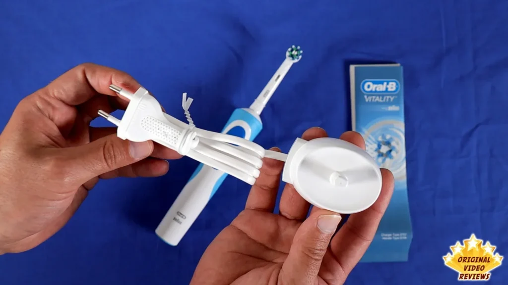 Hands holding the charging unit and its plug next to the toothbrush and retail box on a blue surface
