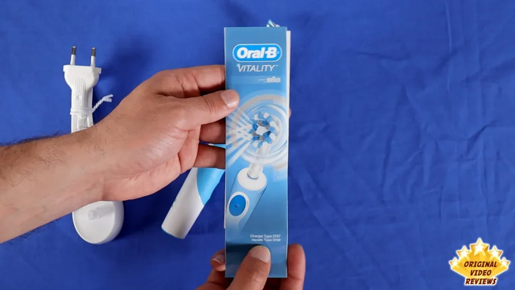 Hand holding the retail box beside the charging unit and the Oral-B Vitality toothbrush on a blue surface